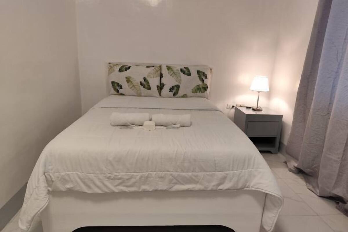 A neatly made bed with white bedding, a headboard with a leaf pattern, a bedside table with a lamp, and a curtain on the right.