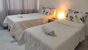 Zwei Einzelbetten mit weißer Bettwäsche und Kissen mit Blättermuster, eine Nachttischlampe und ein Fenster mit Vorhängen.