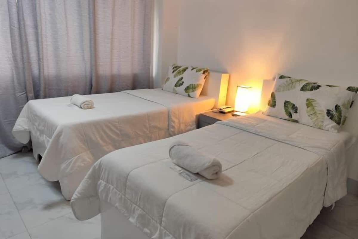 Two single beds with white bedding and leaf-patterned pillows, a bedside lamp, and a window with curtains.
