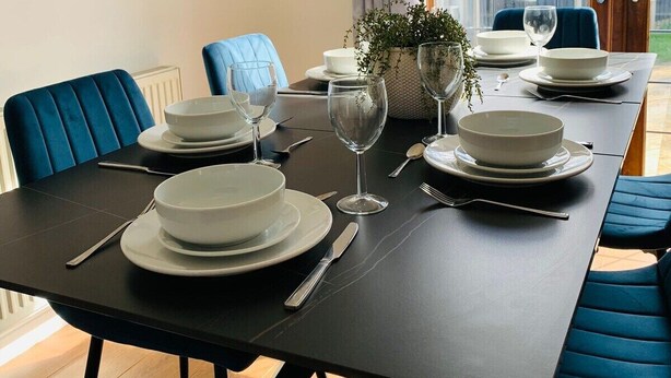 A dining area with a black table set for four, blue chairs, and a view of a garden through a glass door. - Wakefield 3 Bed Home (West Yorkshire)