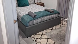 A bedroom with a bed, a nightstand, a lamp, a framed picture on the wall, and a patterned rug. - Home Away From Home (Brierley Hill)