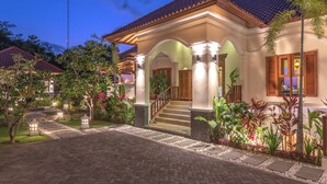 A well-lit, two-story building with a tiled roof, a central staircase, and a landscaped garden with various plants. - Villa Mata Hari (Sukasada)