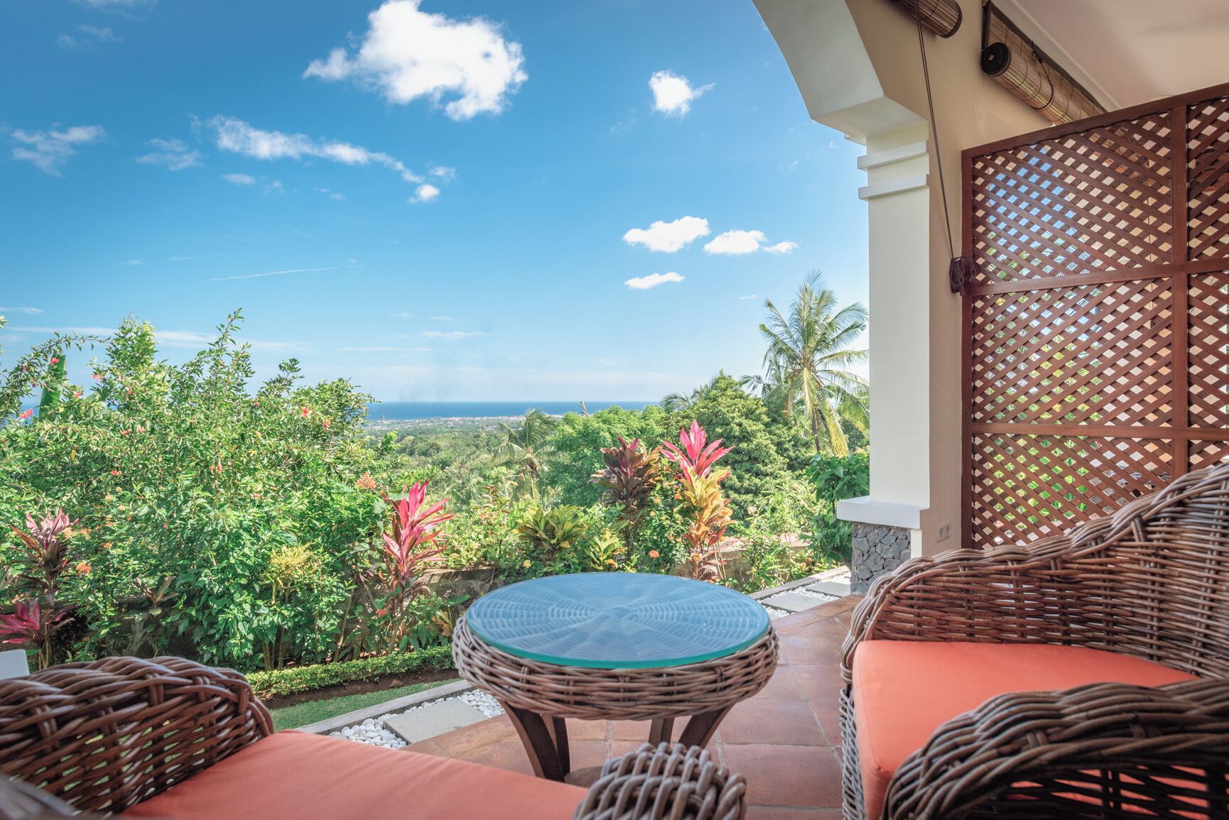 A balcony with wicker furniture, a glass table, and a view of the ocean and greenery.