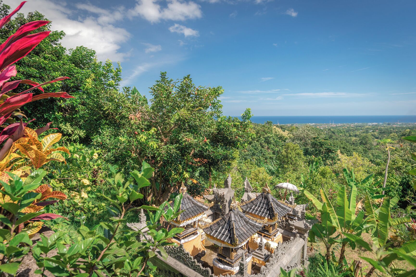 A temple complex with traditional architecture, surrounded by lush greenery and a clear view of the ocean.