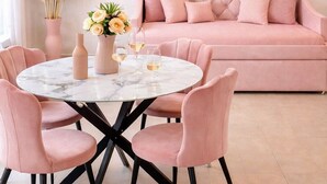 Una sala de estar moderna con un sofá rosa, una mesa redonda de mármol con sillas rosas, y elementos decorativos como un jarrón con flores y un reloj en la pared.