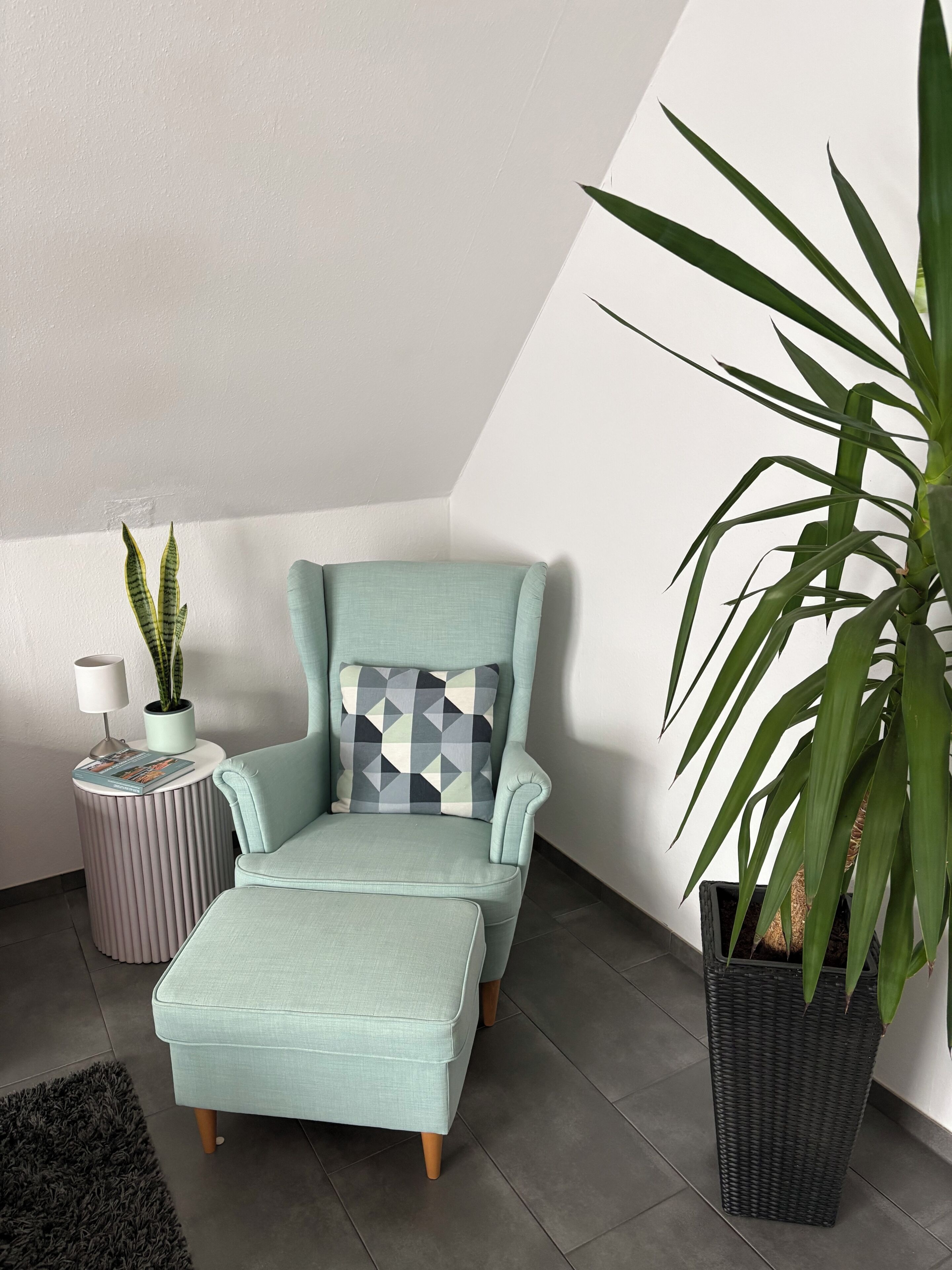 A room with a light blue armchair, a matching ottoman, a patterned pillow, a small side table with a lamp and a plant, and a large potted plant.