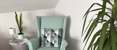 A room with a light blue armchair, a matching ottoman, a patterned pillow, a small side table with a lamp and a plant, and a large potted plant.