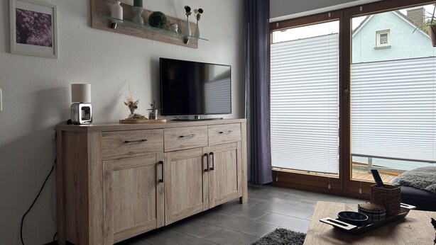 A modern living room with a TV, wooden cabinet, and a sliding glass door to the outside. - Comfortable vacation apartment in the center of Schleiden National Park (Schleiden)