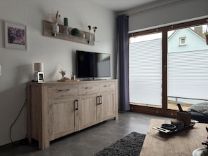 A modern living room with a TV, wooden cabinet, and a sliding glass door to the outside. - Comfortable vacation apartment in the center of Schleiden National Park (Schleiden)