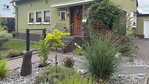 A two-story house with a green exterior, a front porch, and a small garden with various plants and a metal fence. - Comfortable vacation apartment in the center of Schleiden National Park (Schleiden)