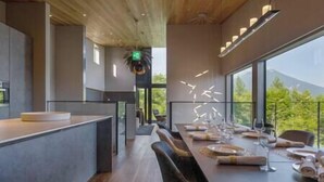 A modern dining area with a long table set for a meal, large windows offering a view of greenery and a mountain, and a contemporary ceiling design. - Rinzan Chalet | 3BR Soga Chalet | Onsen-Style Bath & Yotei View (Rinzan)