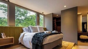 A modern bedroom with a large bed, a nightstand, and a view of trees through the windows. - Rinzan Chalet | 3BR Soga Chalet | Onsen-Style Bath & Yotei View (Rinzan)