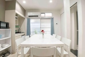 A modern hotel room with a white dining table, white chairs, a small kitchen area, and a large window with curtains. - SunStay Shimbamba I (Tokyo)