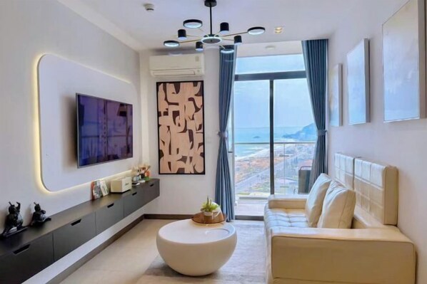 A modern living room with a flat-screen TV, a round coffee table, a sofa, and a large window offering a city view. - Bap's House - CSJ Apartment Vung Tau (Vung Tau)