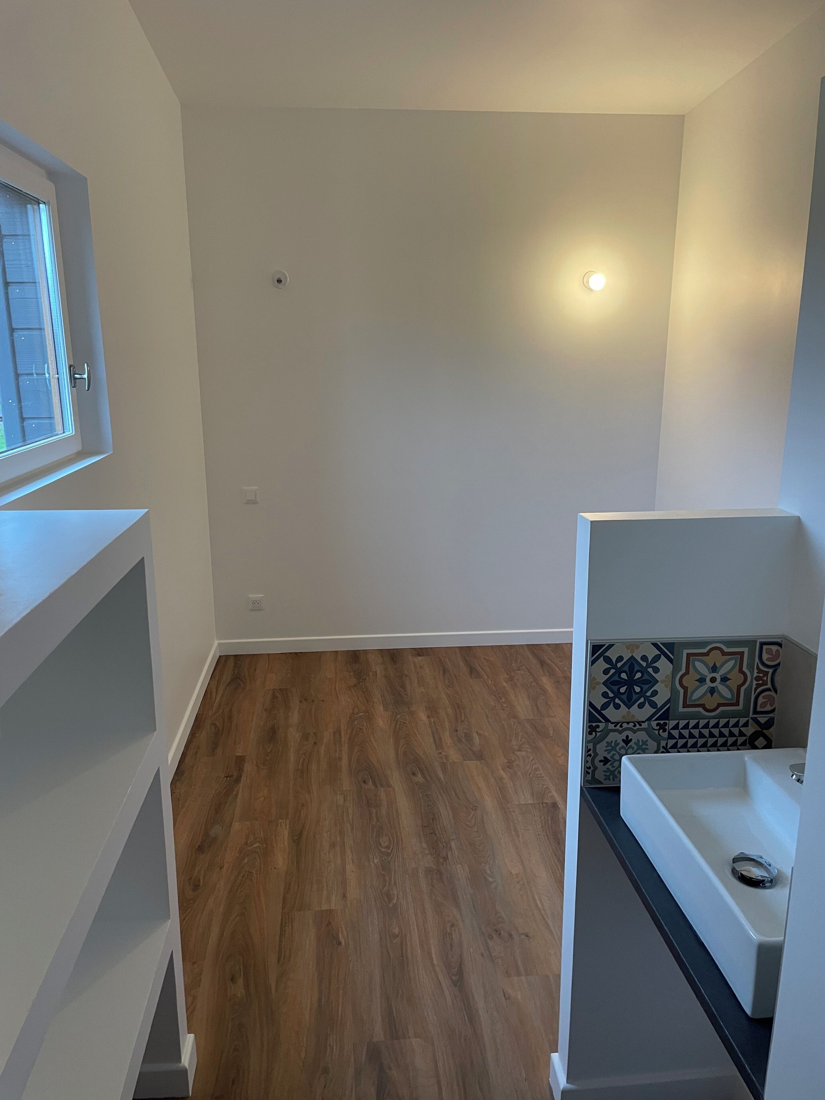 Ein Zimmer mit Holzboden, weißen Wänden, einem Fenster und einer gefliesten Rückwand.