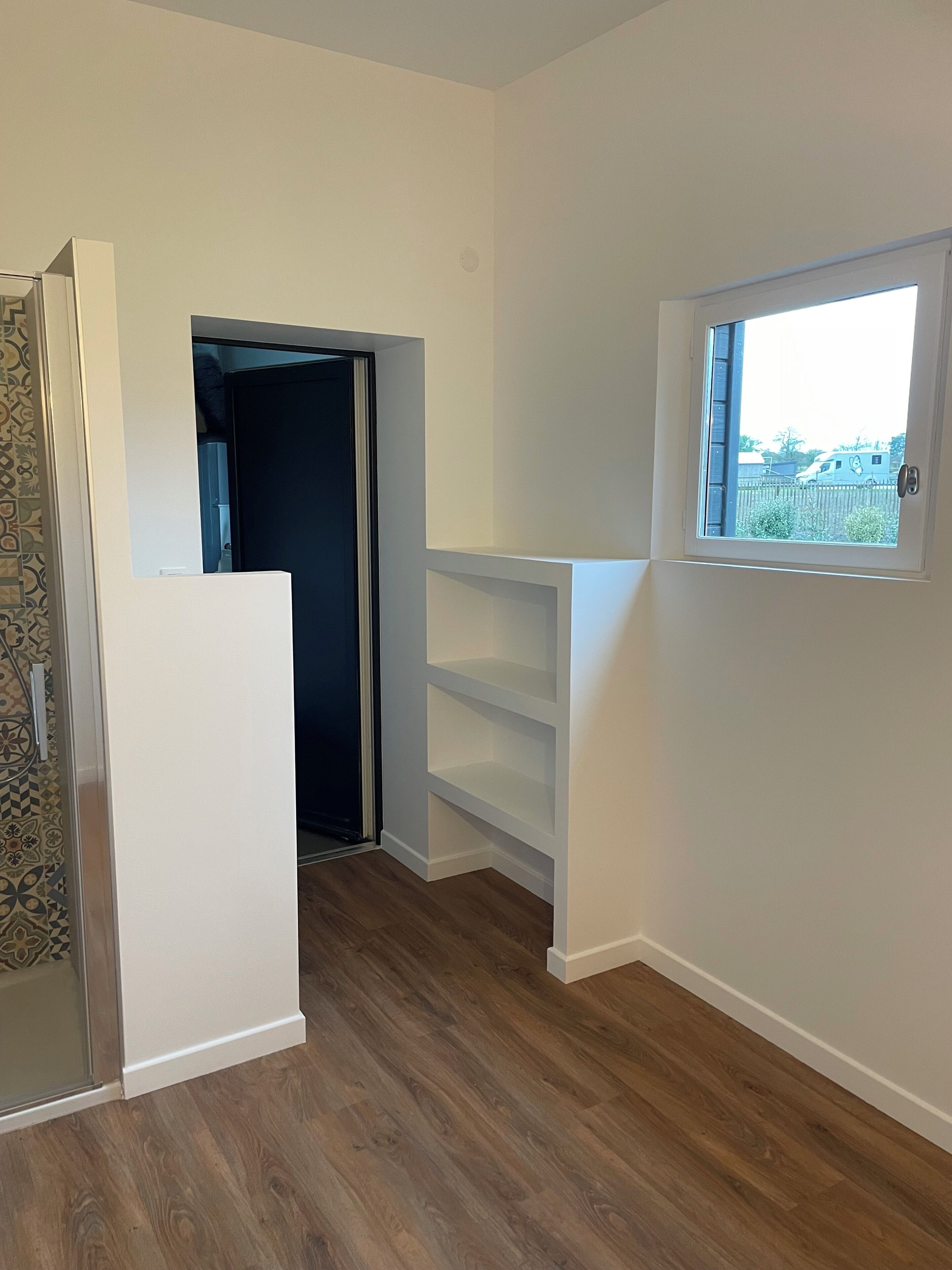 Una habitación con suelo de madera, paredes blancas, una estantería empotrada y ventana con vistas al exterior.
