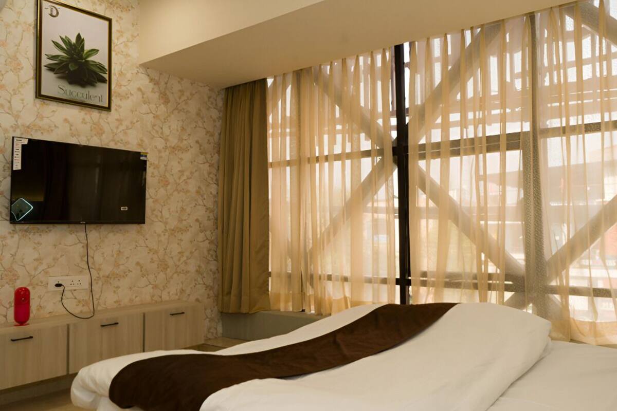 Una habitación de hotel con una cama, un televisor fijado a la pared, una planta y cortinas traslúcidas.