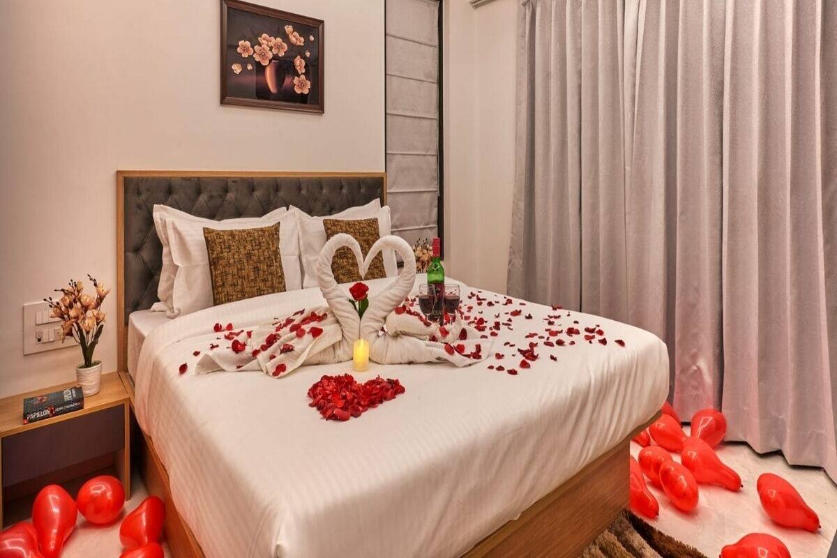 A neatly made bed with rose petals and a heart-shaped decoration, a bedside table with a bottle and a glass, and a large window with curtains.
