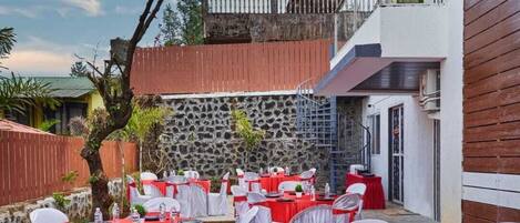 An outdoor dining area with round tables set for a meal, surrounded by chairs with white and red sashes.