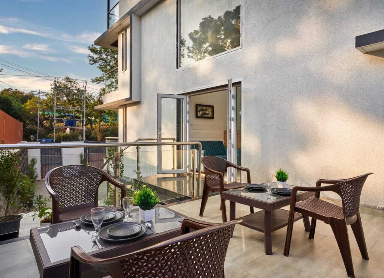 A modern outdoor dining area with a glass table and wicker chairs, a small potted plant, and a view of the building's exterior.