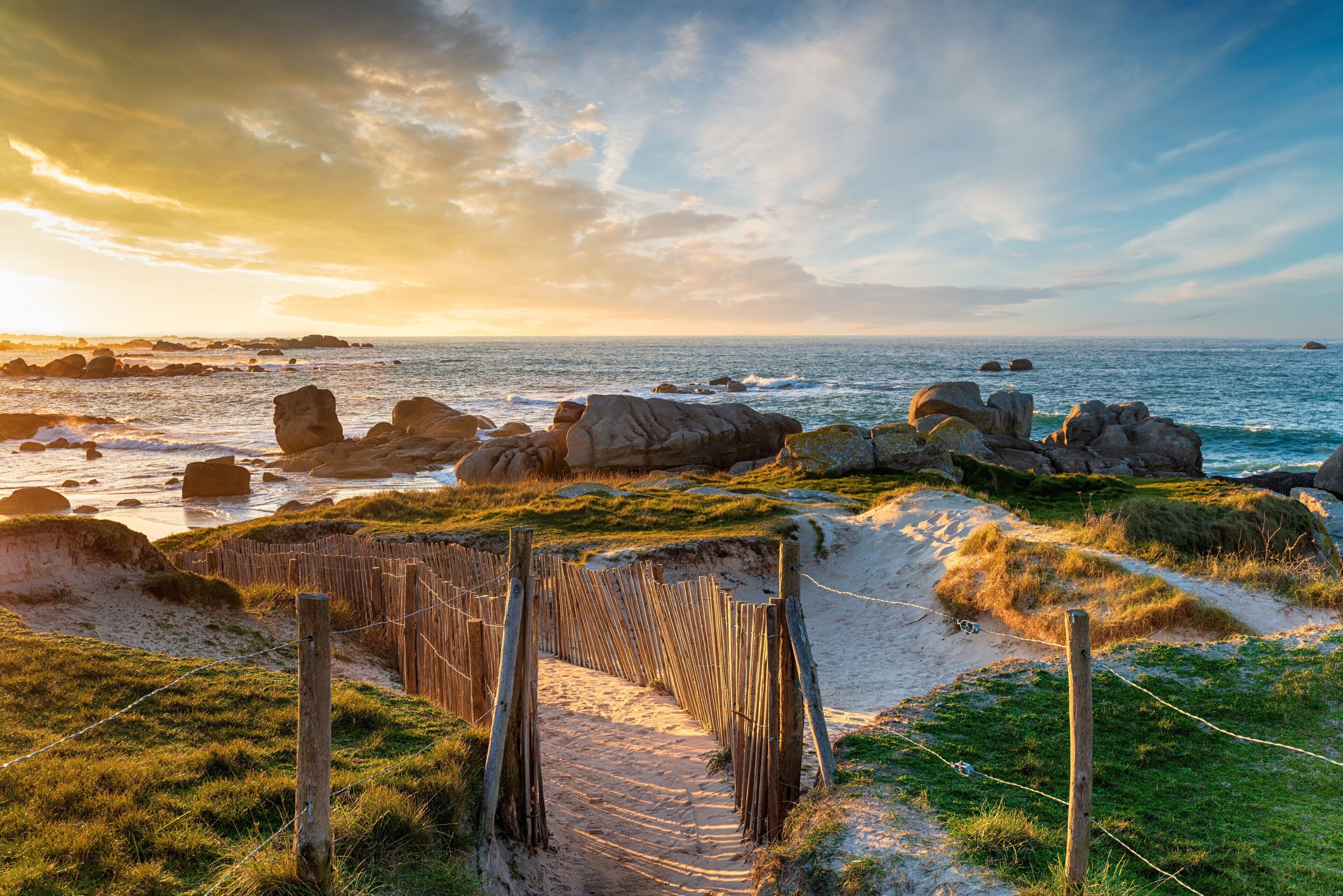 A coastal pathway with a wooden fence, leading to a beach with visible rocks and waves.