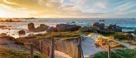 A coastal pathway with a wooden fence, leading to a beach with visible rocks and waves.