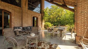 Un patio exterior con mobiliario de mimbre, alfombra de cuero y un edificio de ladrillo con ventanas arqueadas.