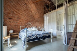 Une pièce avec un lit en métal, un mur en briques et une grande fenêtre avec des rideaux.