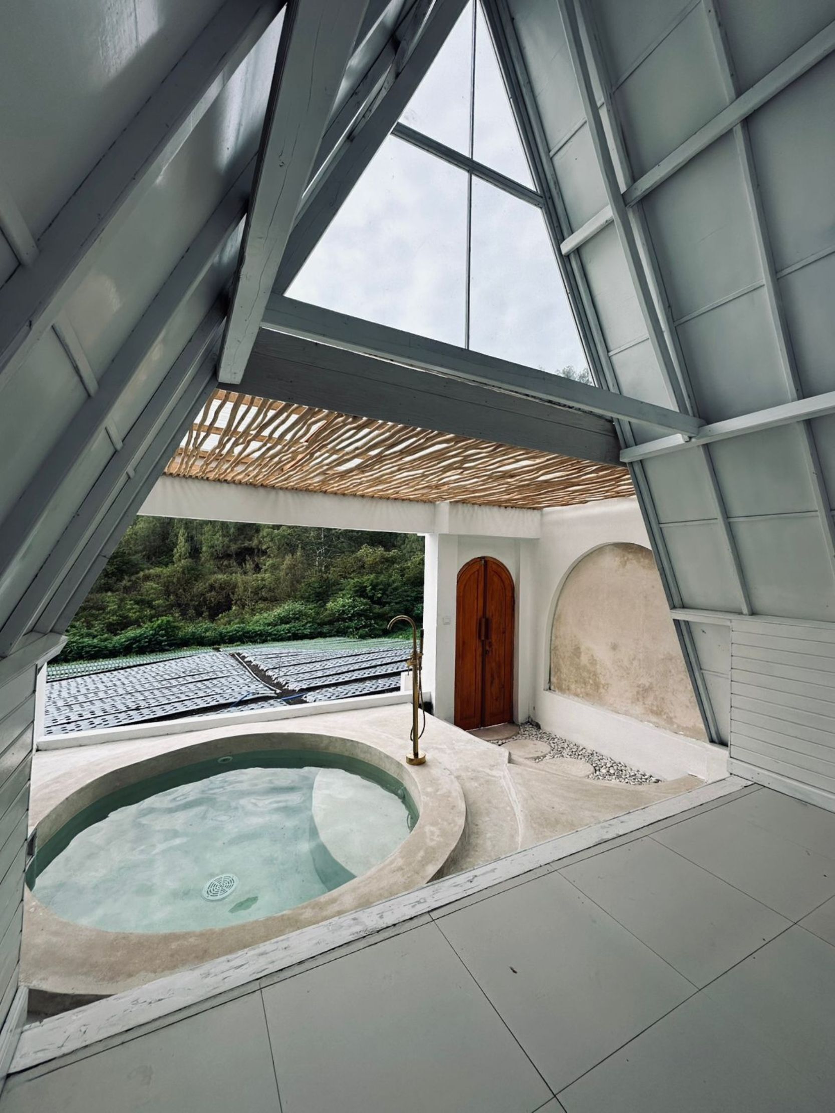 Un espace spa moderne doté d’un jacuzzi, d’une porte en bois et offrant une vue sur un toit à travers une structure en verre.