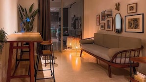 Un espacio de sala acogedor con un sofá, una mesa de centro y un taburete. Hay cuadros en la pared y una planta a la izquierda.