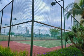 A tennis court enclosed by a netted fence with a red surface, surrounded by greenery and palm trees. - Deluxe 3 Bed 3 Bath Apartment (Johor Bahru)