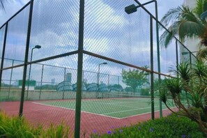 A tennis court enclosed by a chain-link fence with a green and red surface, surrounded by greenery and palm trees.