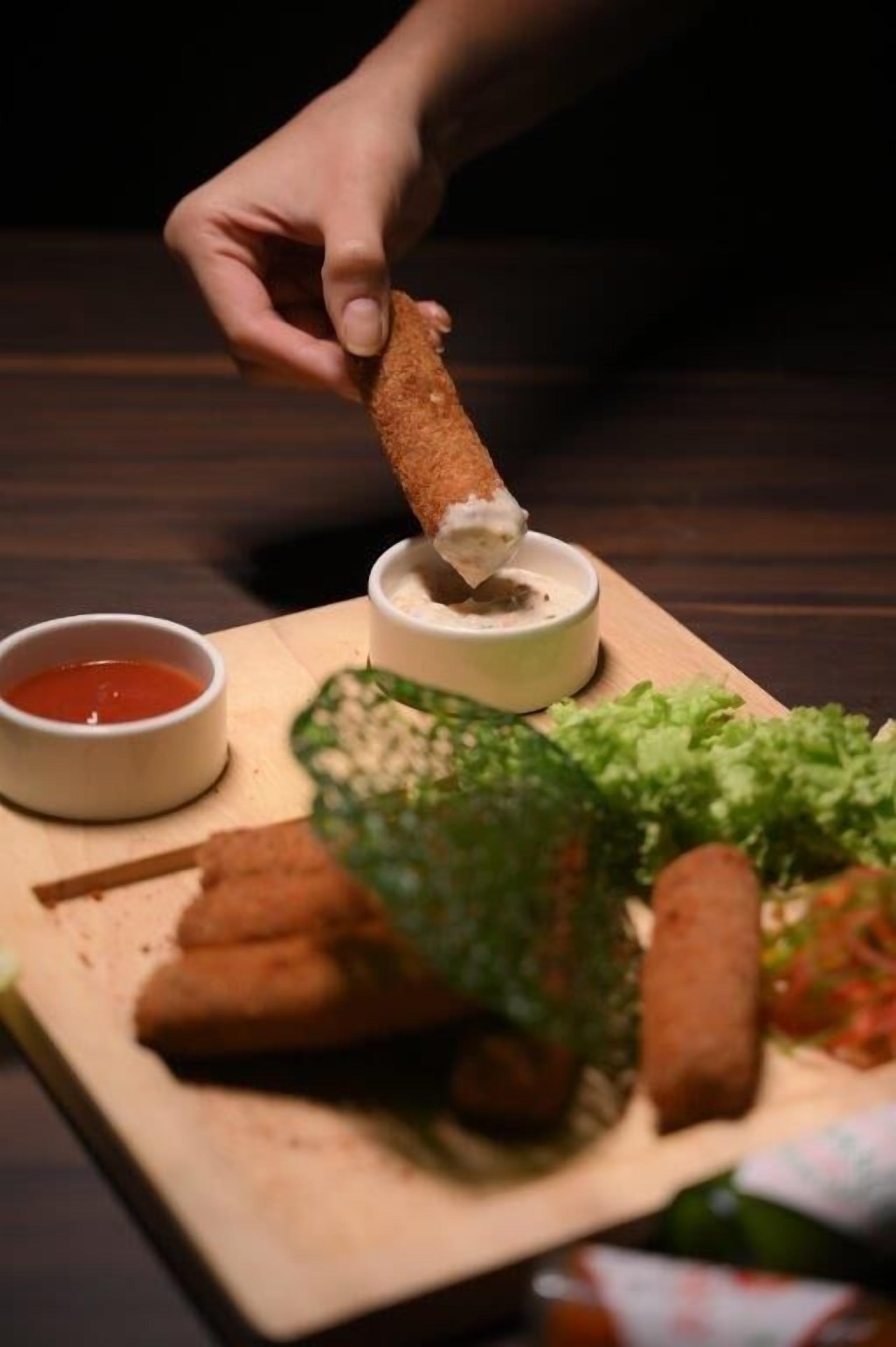 Une main plonge un aliment frit dans un récipient contenant de la sauce, à côté d’un bol de sauce rouge et d’une assiette de légumes verts et d’autres aliments, le tout posé sur une planche en bois.