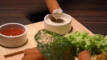 Une main plonge un aliment frit dans un récipient contenant de la sauce, à côté d’un bol de sauce rouge et d’une assiette de légumes verts et d’autres aliments, le tout posé sur une planche en bois.