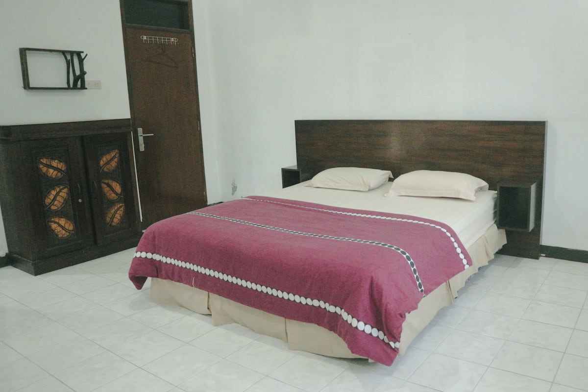 Een bed met een houten hoofdbord, een rood-wit bedovertrek en twee kussens. Links staat een houten garderobe met glazen deuren.