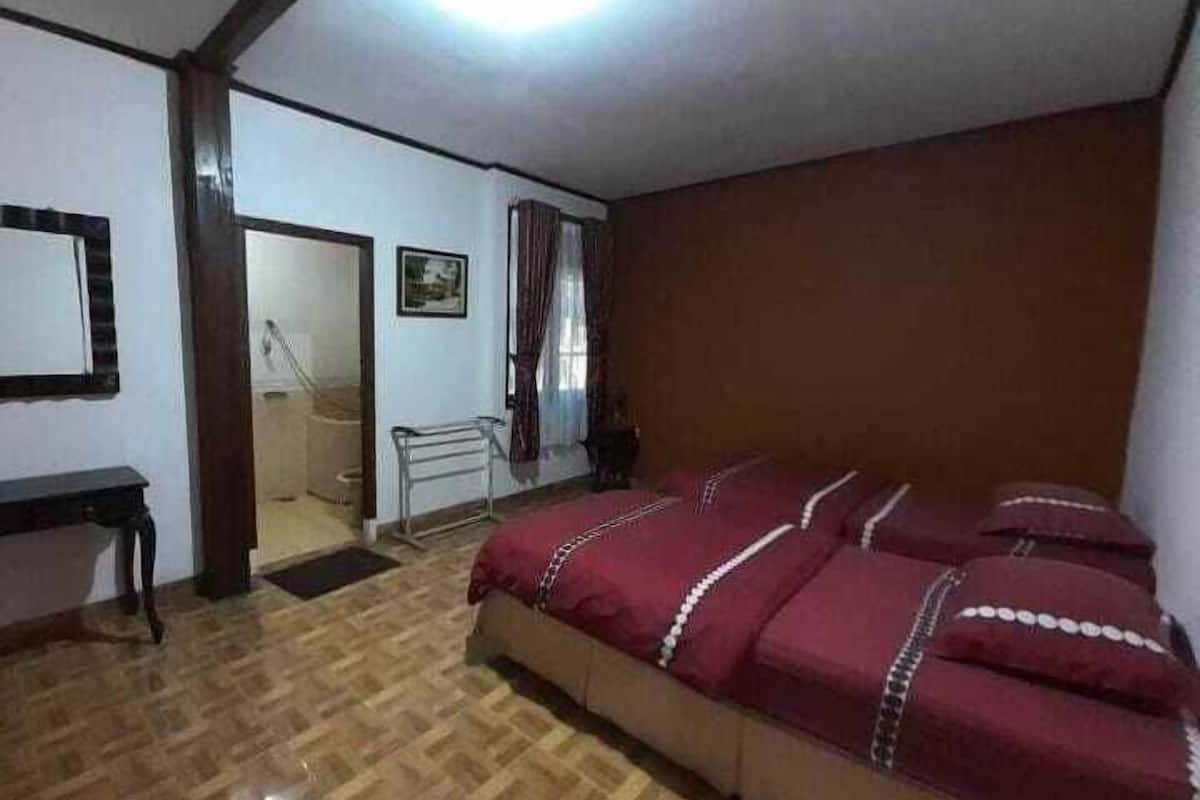 Een slaapkamer met een houten vloer, een bed met een rode sprei, een nachtkastje, een spiegel en een deur naar een andere kamer.