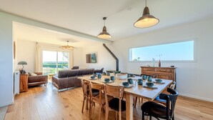 Een ruime woonkamer met een houten eettafel, stoelen en een bank. Er is een keuken met kastjes en een raam.