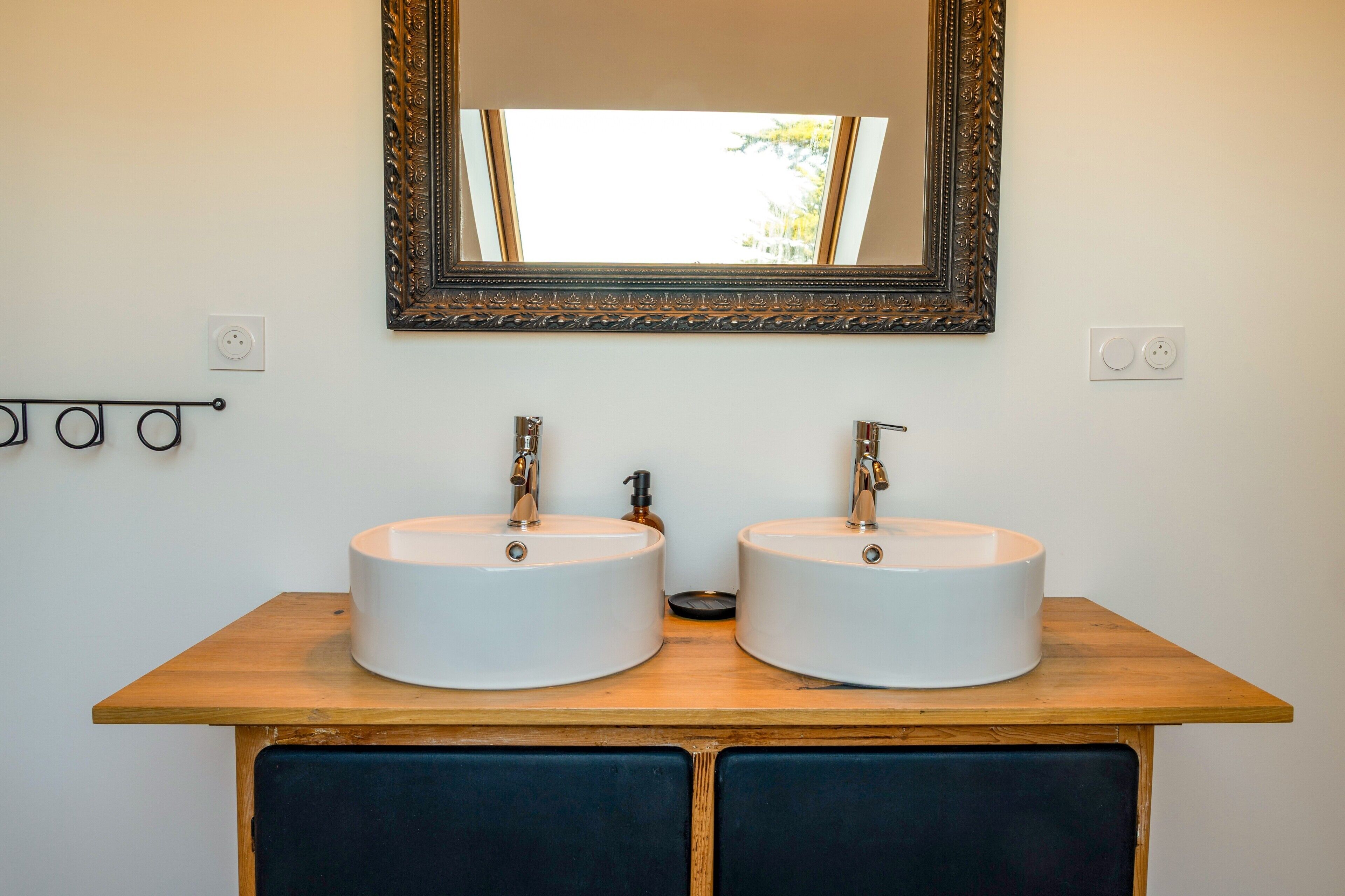 A bathroom with two white sinks, a wooden vanity, a mirror with an ornate frame, and a skylight.