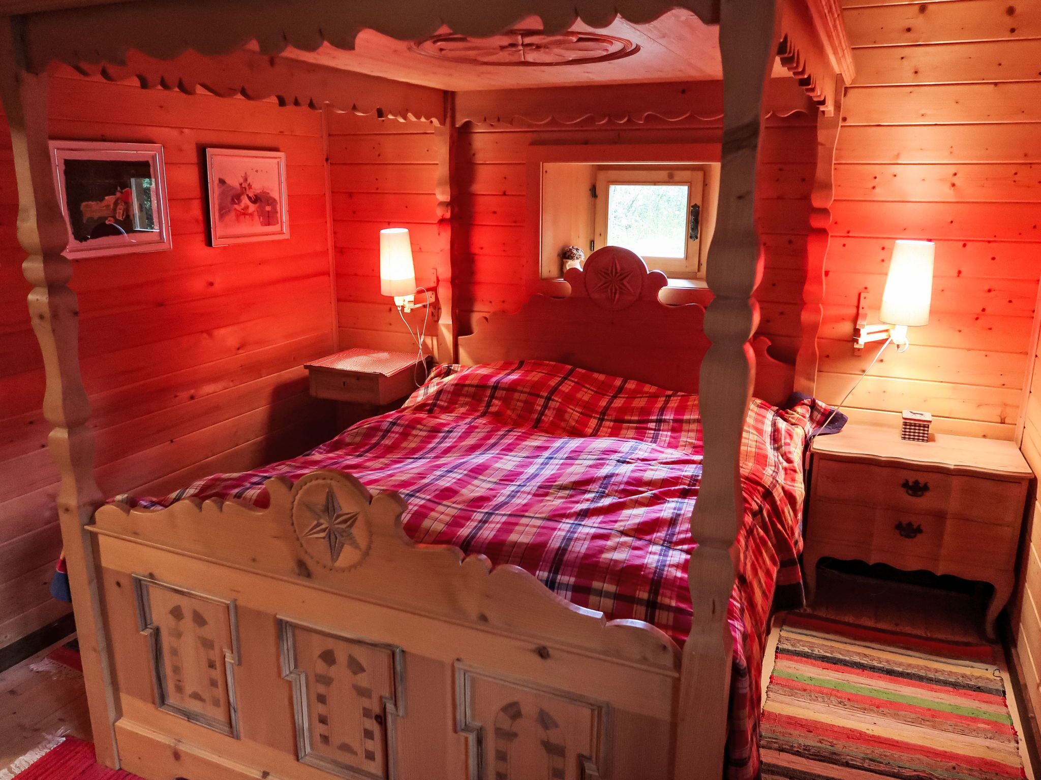 Un lit à baldaquin en bois, recouvert d’une couverture à carreaux, deux tables de chevet avec des lampes, une fenêtre donnant sur un beau paysage et une petite commode en bois.