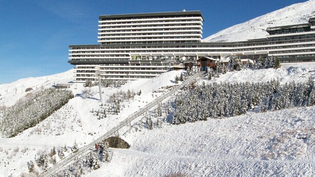 Un hôtel moderne à plusieurs étages, situé sur une colline enneigée, avec un télésiège visible.