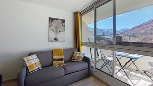 Un canapé gris agrémenté de coussins à motifs, une œuvre d’art encadrée représentant le visage d’un loup, et une vue sur un paysage montagneux par de grandes fenêtres.