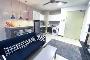 A modern living room with a sofa, a coffee table, and a ceiling fan. There is a kitchen area with a washing machine and cabinets. - KL Gateway Residence (Kuala Lumpur)