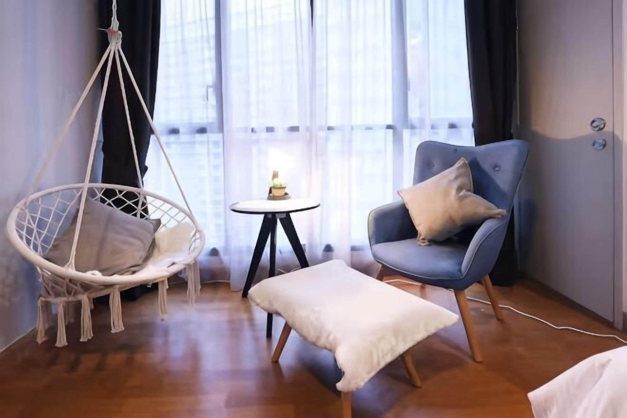 A room with a hanging chair, a small round table, a blue armchair with a pillow, and a white ottoman.