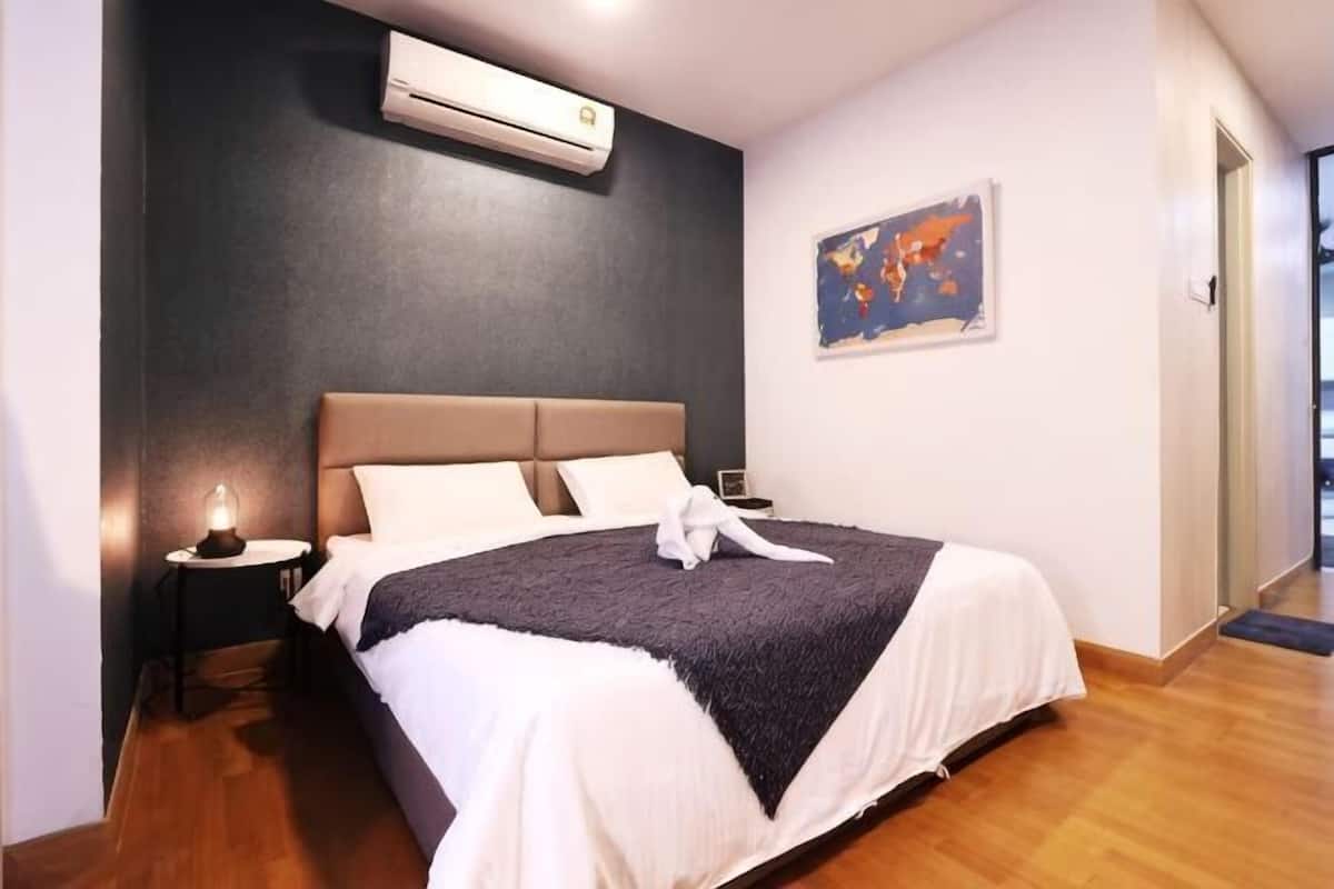 A bedroom with a bed, an air conditioner, a nightstand, a painting, and a wooden floor.
