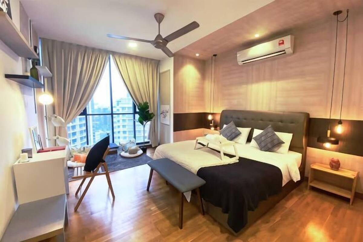 A modern hotel room with a large bed, a desk, a chair, a bench, a ceiling fan, and a view of the city.