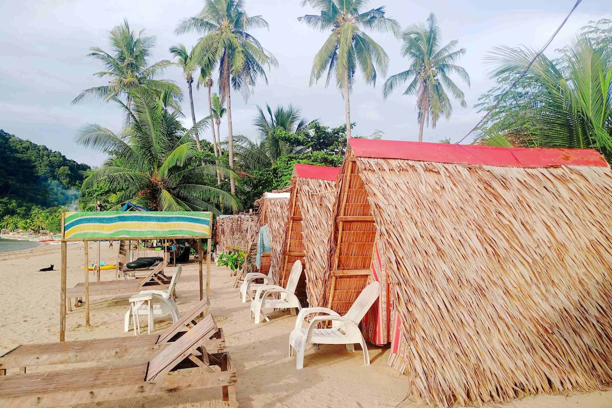 Une plage avec des cabanes à toit de chaume, des chaises en plastique blanches et un parasol rayé.