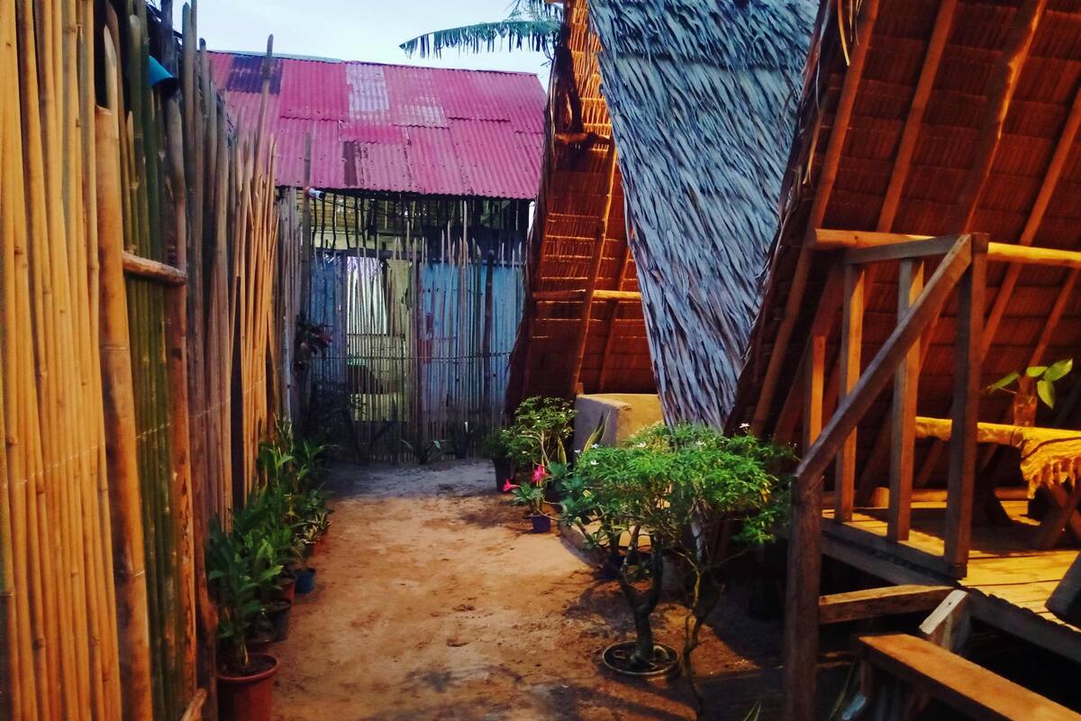 Un sentier étroit bordé de clôtures en bambou et de constructions en bois, qui mène à un bâtiment au toit de chaume.