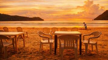 Un entorno de playa con mesas y sillas dispuestas para comer, con vistas al mar y una puesta de sol al fondo.