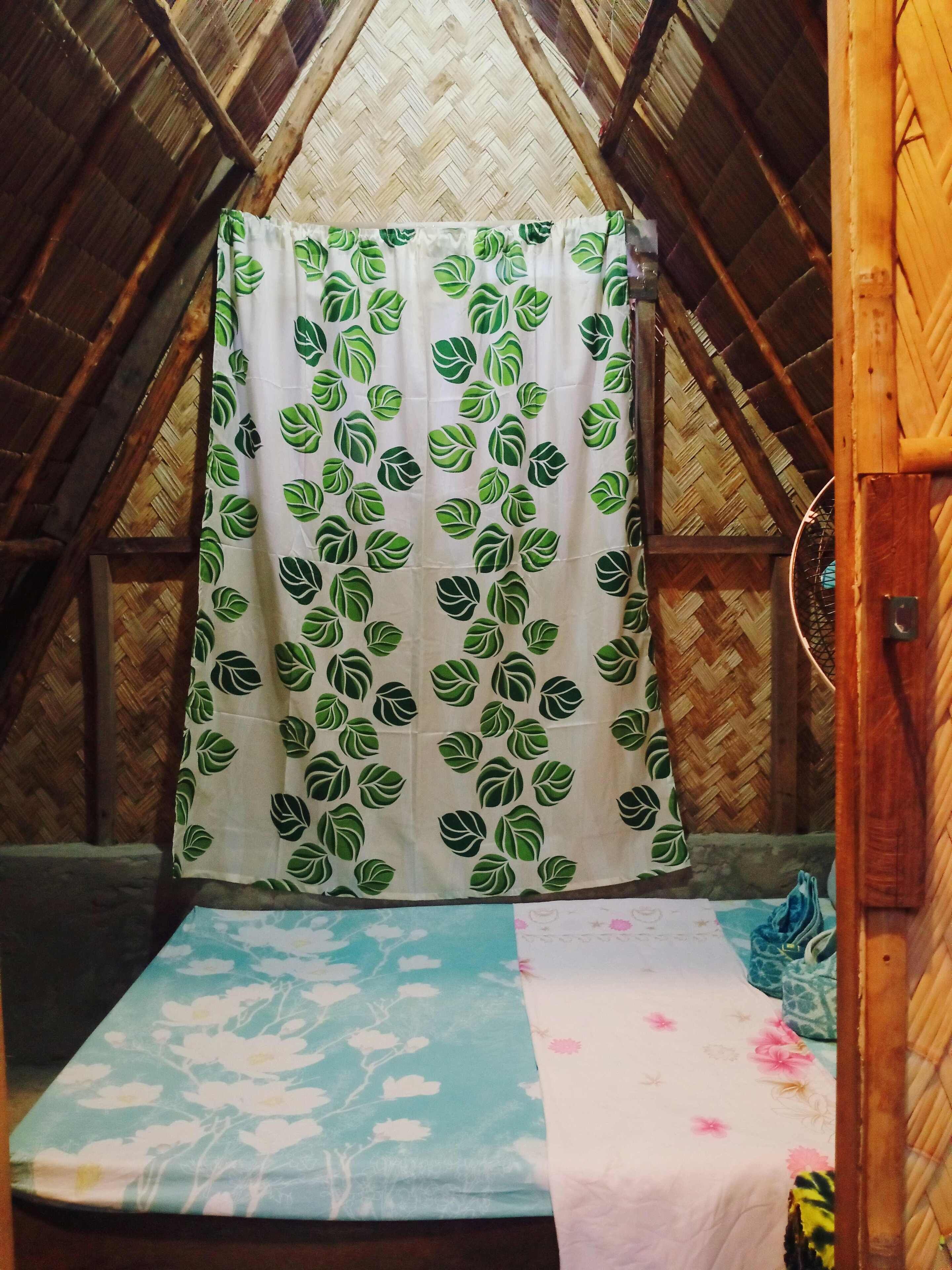 A small, cozy room with a bed covered in a leaf-patterned curtain, a blue and white floral bedspread, and a wooden ceiling.