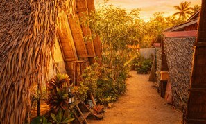 A narrow pathway flanked by thatched-roof structures and lush greenery, suggesting a rustic, tropical setting. - Amwani Beach Camp (El Nido)